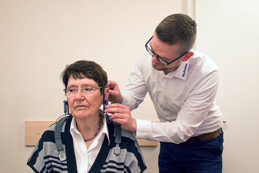 Ein Hörakustiker passt einer älteren Frau ein Hörgerät zur Hörtest-Vorbereitung an.
